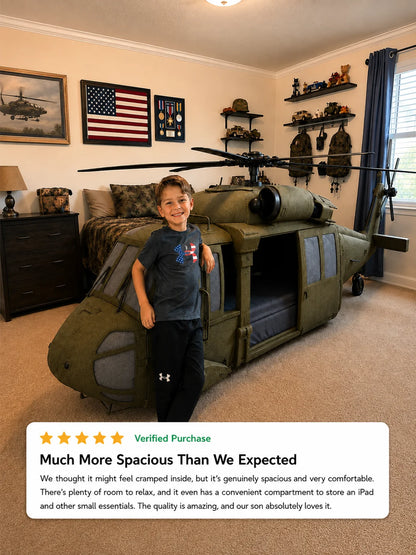 A young boy stands smiling next to a large olive green military helicopter bed in a spacious, military-themed bedroom. The room features a bed with camouflage bedding, military wall decorations, and shelves with gear.